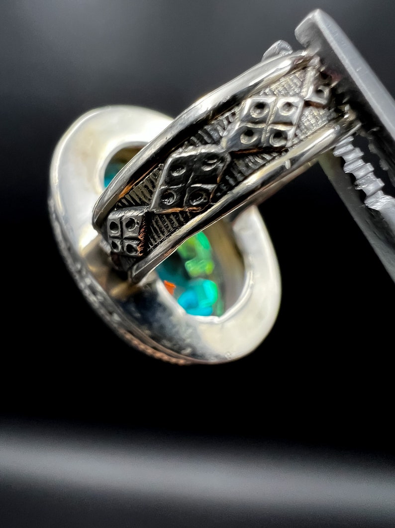 May include: Close-up of a silver ring with an oval-shaped, iridescent gemstone. The ring's band features a detailed, geometric pattern. The gemstone displays a vibrant play of colors, including blues, greens, and oranges, against a dark background.