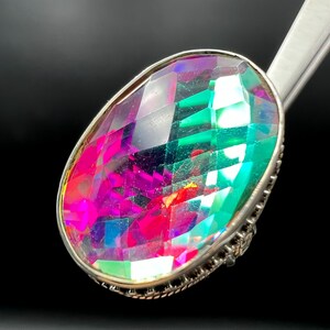 May include: A large, oval-cut gemstone ring with a silver-colored setting. The gemstone displays a vibrant array of colors, including pink, green, and purple, with a faceted surface that catches the light. The ring is set against a dark background.
