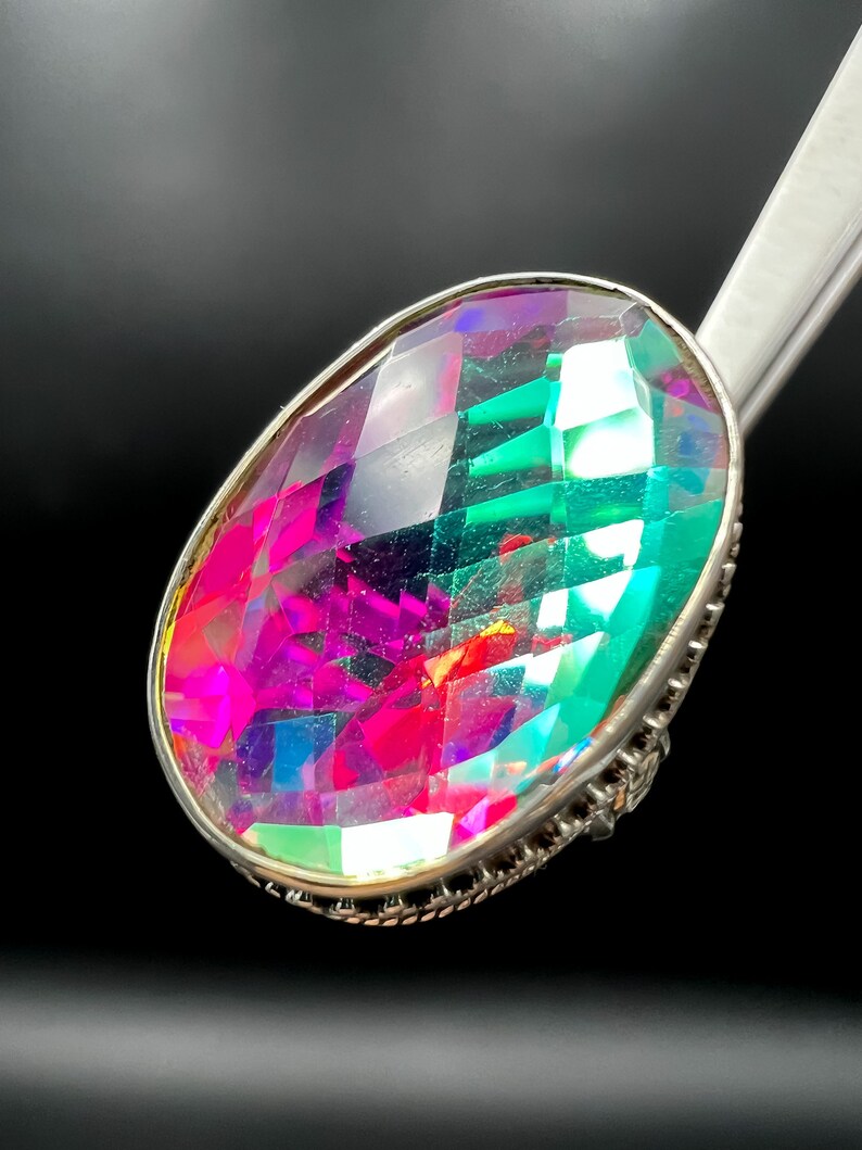 May include: A large, oval-cut gemstone ring with a silver-colored setting. The gemstone displays a vibrant array of colors, including pink, green, and purple, with a faceted surface that catches the light. The ring is set against a dark background.