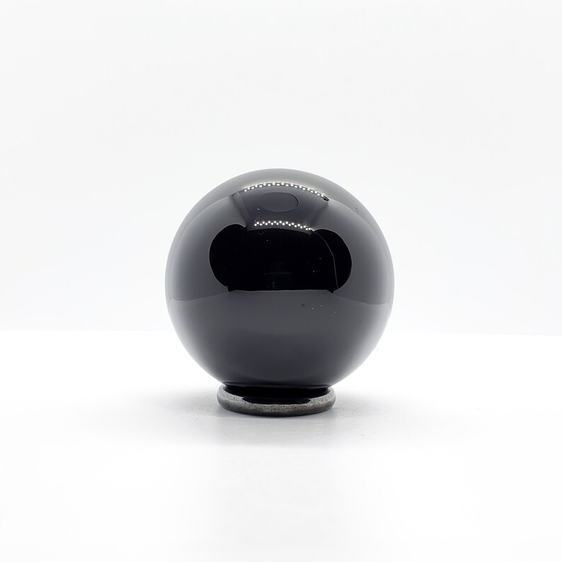 Op de afbeelding: Een zwarte obsidiaans bol op een kleine metalen standaard. De bol is hoogglanz gepolijst en reflecteert licht.