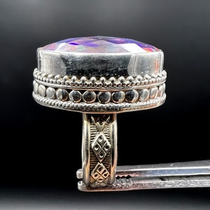 May include: A silver ring with a large, iridescent, oval-shaped gemstone. The ring has a detailed band with an ornate design and a beaded border around the gemstone. The gemstone displays a range of colors, including purple, blue, and green.