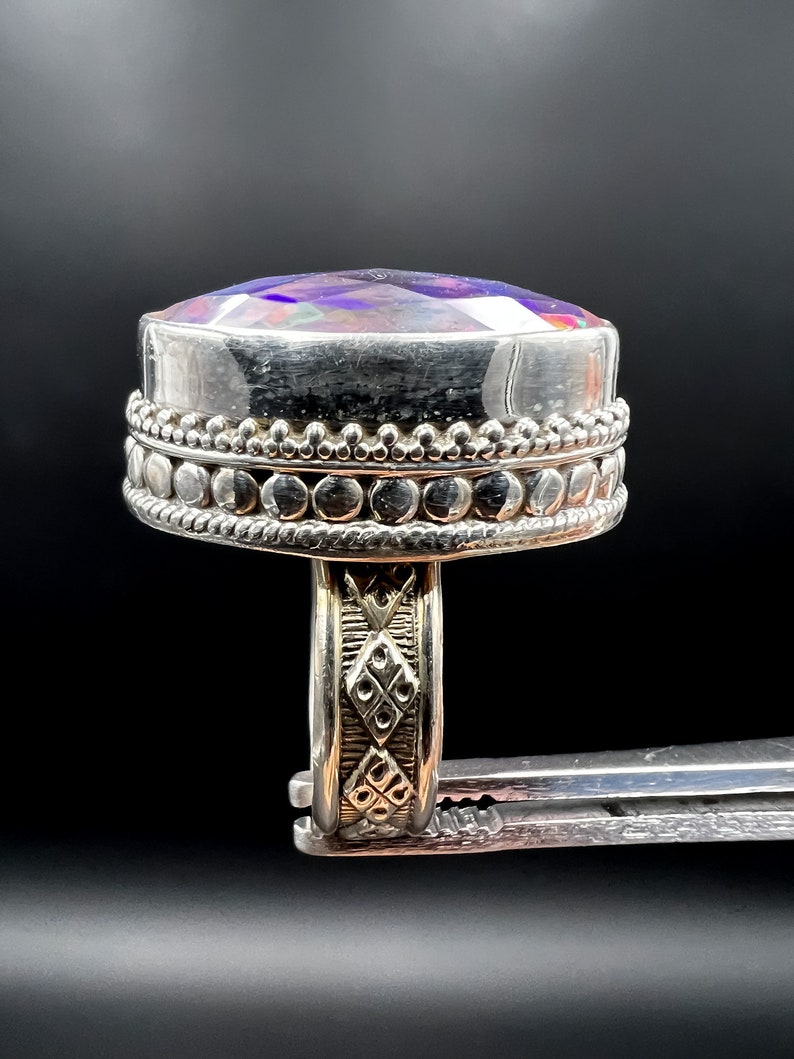 May include: A silver ring with a large, iridescent, oval-shaped gemstone. The ring has a detailed band with an ornate design and a beaded border around the gemstone. The gemstone displays a range of colors, including purple, blue, and green.
