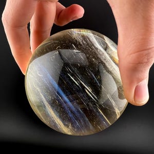 May include: A clear, spherical crystal ball with internal streaks of gold, white, and blue. The crystal is held in a hand against a black background. The ball has a polished, reflective surface.