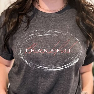 May include: A dark heather grey t-shirt with a white circle graphic and the word "THANKFUL" written in pink inside the circle.
