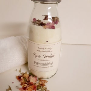 Könnte beinhalten: Ein Glasgefäß gefüllt mit einem weißen Badezusatz und getrockneten Rosenblüten. Das Gefäß hat einen silbernen Deckel und ein Etikett mit der Aufschrift "Beauty & Soap Seifenmanufaktur Rose Garden mit Kakaobutter Buttermilchbad www.beautyandsoap.de 150g".