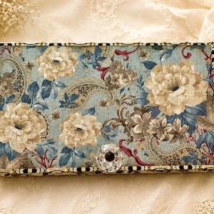 May include: Rectangular decorative box with a floral pattern in shades of blue, beige, and brown. The box features a paisley design and a crystal knob. The edges are trimmed with gold and black accents.