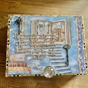 May include: A rectangular decorative box featuring a blue sky background with a staircase leading to an open book illustration. The cover includes two metal skeleton keys, a stack of books, and cursive Italian text. A clear crystal knob is attached.