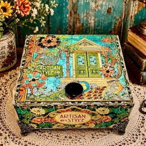 May include: A decorative box with a colorful, ornate design featuring a peacock, flowers, and a house facade. The box has a black knob on top and the words "ARTISAN STYLE" are displayed on the front. The box is resting on a lace doily.