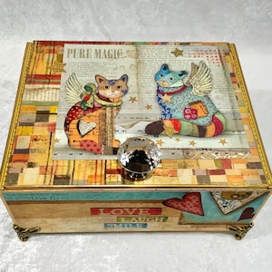 May include: A rectangular decorative box featuring a collage design with two winged cats, the text PURE MAGIC, and a faceted crystal knob. The front panel displays the words LOVE, LAUGH, and SMILE, with heart motifs and a warm, multi-colored pattern.