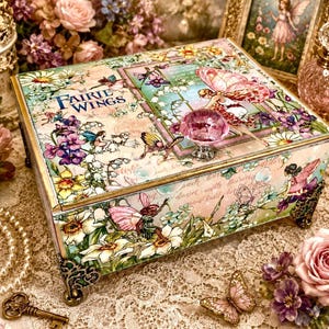 May include: A decorative wooden box with a floral and fairy design. The box features the words "FAIRIE WINGS" and a pink crystal knob. The box is adorned with images of fairies, butterflies, and flowers. The box has ornate metal feet.
