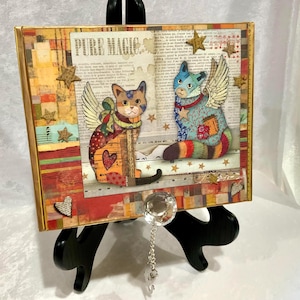 May include: A framed mixed-media artwork featuring two whimsical cat figures with wings, set against a backdrop of vintage paper and star accents. The artwork has a gold frame and the words "PURE MAGIC" are visible.