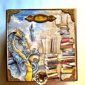 May include: A wooden box with a dragon and a stack of books on the front. The dragon is blue and gold and is holding a glowing orb. The books are stacked on top of each other and are bound in different colors. The box is decorated with gold trim and has a small, clear crystal on the front.