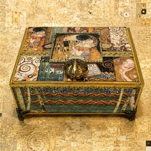 May include: A rectangular decorative box featuring a gold-toned metal frame and feet. The exterior surfaces are adorned with colorful, intricate art prints inspired by Gustav Klimt, including figures and patterns. A round, gold-toned metal knob sits on top.