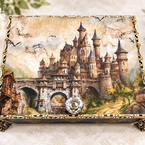 May include: A rectangular decorative box featuring a detailed illustration of a stone castle with multiple towers and a bridge on its lid. The box has a clear, faceted crystal knob handle and ornate, metallic gold-toned feet at each corner.