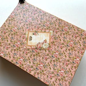 May include: A decorative box with a pink floral pattern featuring roses and other flowers. The box has metal corner protectors and a rectangular label with a crown and floral accents.