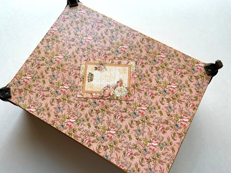 May include: A decorative box with a pink floral pattern featuring roses and other flowers. The box has metal corner protectors and a rectangular label with a crown and floral accents.