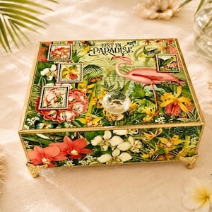 May include: A decorative box with a tropical theme. The box features a flamingo, flowers, and the text "Lost in Paradise." It has a clear glass knob and gold-colored feet.