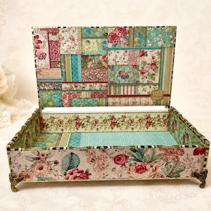 May include: A rectangular decorative box with an open lid, featuring a patchwork floral pattern in shades of pink, teal, and cream. The interior and exterior surfaces are covered in paper with rose motifs. It stands on four small ornate metal feet.