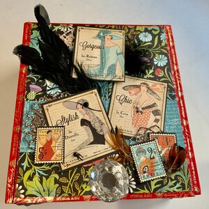 May include: A vintage-style scrapbook cover with a floral design and several paper embellishments. The embellishments include images of women in vintage clothing, text that reads "Gorgeous", "Stylish", "Chic", and "Van Leef", and a few small paper stamps.