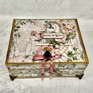 May include: A decorative box with a floral and script design, featuring the text "HOUSE OF ROSES." The box has a gold trim, a pink bow, and decorative feet. The box is adorned with roses and a bird silhouette.
