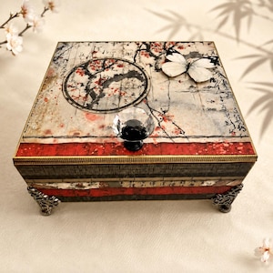 May include: A square decorative box featuring a top lid with a black and white butterfly, a circular ink-wash style tree illustration, and red accents. The box has a clear faceted crystal knob, dark textured sides with red bands, and four ornate metal feet.