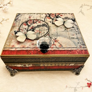 May include: A square decorative box with a Japanese-inspired design. The box features a red and gold border, a black knob, and two white butterflies. The top has a map-like design with red floral accents and a black circle.
