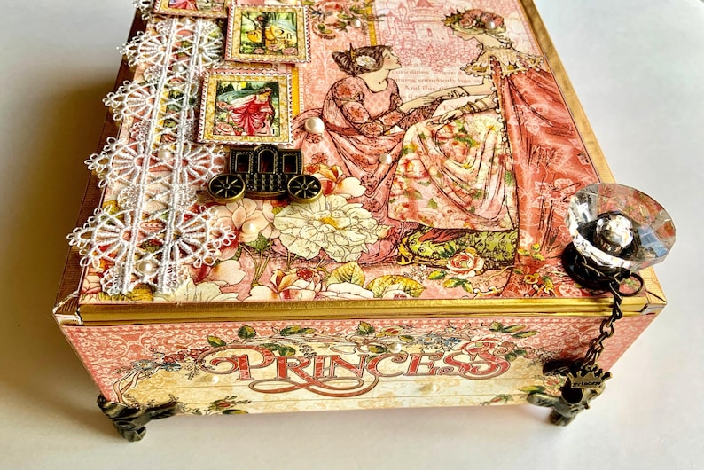 May include: A decorative box with a princess theme. The box features a pink floral design, lace trim, and a crystal knob. The word "PRINCESS" is displayed on the front. Decorative stamps and a carriage adorn the top.