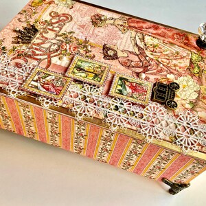 May include: A decorative box with a pink and floral design. The box features a white lace trim, decorative stamps, and a crystal knob. The sides have a pink and yellow striped pattern. The box is supported by decorative metal feet.