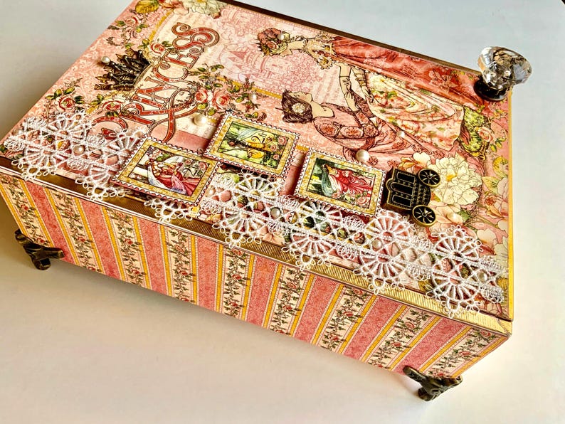 May include: A decorative box with a pink and floral design. The box features a white lace trim, decorative stamps, and a crystal knob. The sides have a pink and yellow striped pattern. The box is supported by decorative metal feet.