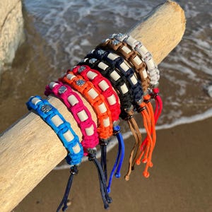 May include: A collection of six paracord bracelets featuring woven nylon cords in blue, pink, orange, black, brown, and grey. Each bracelet incorporates white rectangular beads and a small silver-toned metal charm, finished with adjustable cord ends.