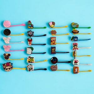 May include: A set of 16 colorful hair clips with various designs, including food, cartoon characters, and geometric shapes. The clips are attached to yellow, pink, and blue hair ties.