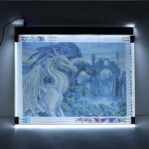 May include: A white LED light pad with a black frame and clips to hold a diamond painting in place. The diamond painting depicts two dragons in a blue and white landscape.