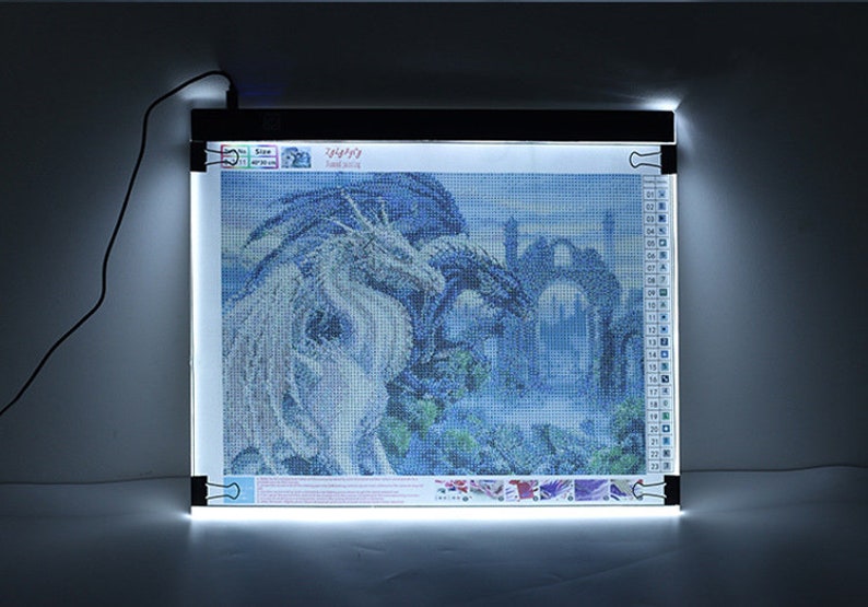 May include: A white LED light pad with a black frame and clips to hold a diamond painting in place. The diamond painting depicts two dragons in a blue and white landscape.