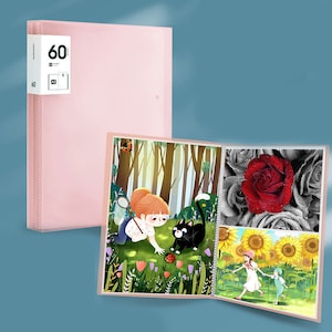 May include: A pink photo album with a clear plastic cover. The album is open to reveal three pages with colorful illustrations. The first page shows a girl with a butterfly net chasing a black cat in a forest. The second page shows a red rose with water droplets on the petals. The third page shows two people walking in a field of sunflowers.