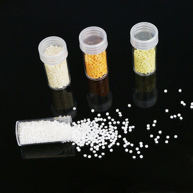 May include: Three small clear plastic jars filled with white, yellow, and orange round beads. A fourth jar is tipped over, spilling white beads onto a black surface.