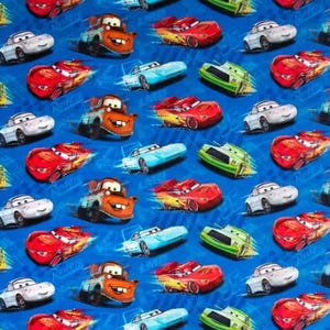 May include: A blue fabric featuring a repeating pattern of animated cars in various colors. The cars include red, white, orange, green, and blue vehicles. The word "Rusteze" is visible on some of the cars.