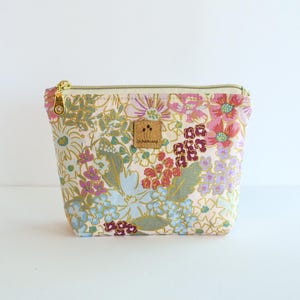 May include: A zippered pouch featuring a light pink background with a multi-colored floral pattern in shades of blue, green, red, and purple. A small square cork label with the text DUNSINANE is centered on the front, and a gold metal zipper.