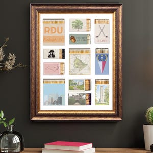 May include: A framed artwork featuring a collection of matchboxes with various designs. The matchboxes display images of a tree, an acorn, a map, and the North Carolina state outline. The artwork is in a brown wooden frame.