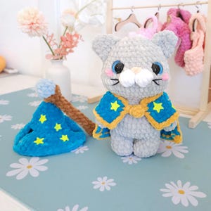 May include: A handmade gray cat plushie with blue eyes, pink cheeks, and a white muzzle, wearing a blue and yellow cape with yellow star accents. A matching blue wizard hat with yellow stars and a brown wand are also present.