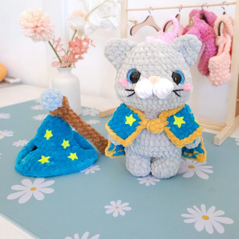 May include: A handmade gray cat plushie with blue eyes, pink cheeks, and a white muzzle, wearing a blue and yellow cape with yellow star accents. A matching blue wizard hat with yellow stars and a brown wand are also present.
