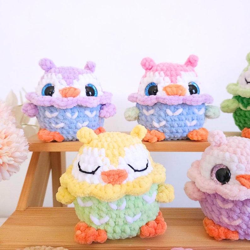 Pu&ograve; includere: Una collezione di figurine di gufo all'uncinetto fatte a mano in vari colori. I gufi presentano corpi blu, viola, rosa e gialli con colori contrastanti per le ali e la testa. Ogni gufo ha grandi occhi scintillanti e piedi arancioni.