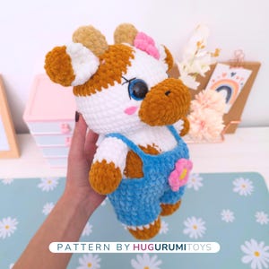 May include: A handmade crochet cow plushie with white and brown fur, wearing blue overalls with a pink flower. The cow has large blue eyes, pink cheeks, and a pink bow. The text "PATTERN BY HUGURUMITOYS" is at the bottom.