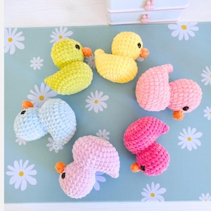 Puede incluir: Una colecci&oacute;n de patitos de ganchillo hechos a mano en varios colores: amarillo, azul claro, rosa y fucsia. Cada patito tiene un pico naranja y ojos negros. Los patitos est&aacute;n dispuestos sobre una superficie azul claro con patrones de margaritas blancas.