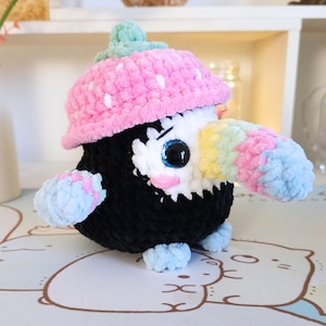 May include: A crocheted toucan plush toy wearing a pink and white polka dot hat. The toucan has a black body, a white face, and a rainbow-coloured beak.