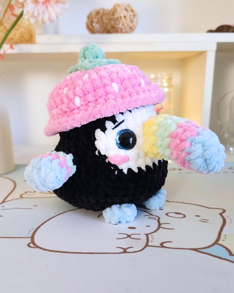 May include: A crocheted toucan plush toy wearing a pink and white polka dot hat. The toucan has a black body, a white face, and a rainbow-coloured beak.