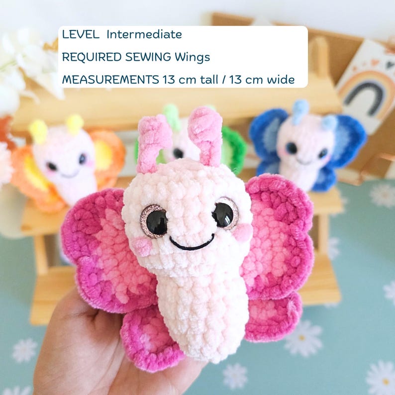 Puede incluir: Primer plano de un peluche de mariposa rosa y fucsia hecho a mano, de 13 cm de alto y 13 cm de ancho, con ojos grandes y brillantes y una cara sonriente. Se ven otros peluches de mariposa de colores. Texto en la imagen: NIVEL Intermedio, REQUIERE COSER Alas.