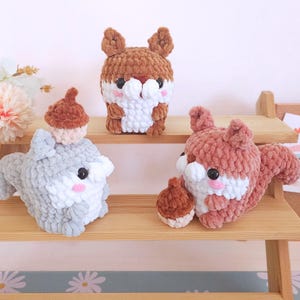 Op de afbeelding: Drie handgemaakte gehaakte eekhoornknuffels in bruine, grijze en roze tinten. Elke knuffel heeft een witte buik, zwarte ogen en roze wangen. E&eacute;n eekhoorn houdt een eikel vast, een andere draagt een eikelhoed en de derde staat rechtop.