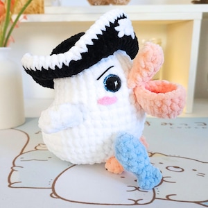 May include: A white crocheted stuffed toy bird wearing a black and white crocheted pirate hat. The bird has a pink crocheted beak and blue crocheted fish in its hand.