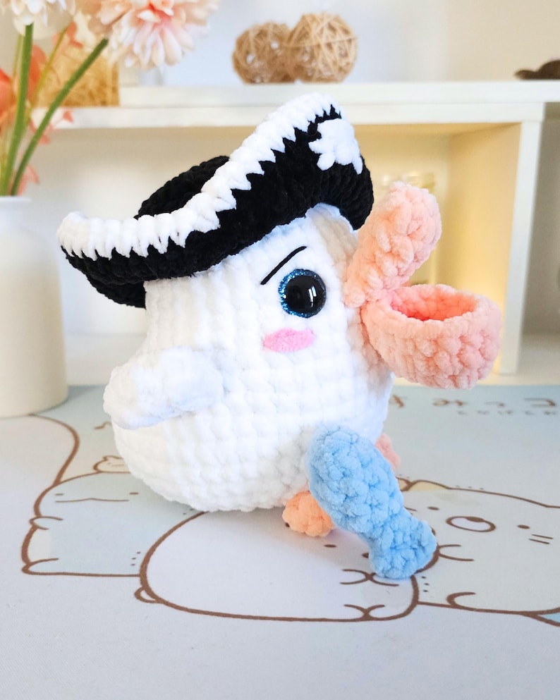 May include: A white crocheted stuffed toy bird wearing a black and white crocheted pirate hat. The bird has a pink crocheted beak and blue crocheted fish in its hand.