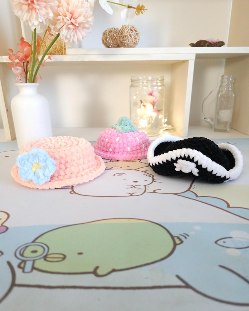 May include: Three crocheted hats in pink, blue and black. The pink hat has white polka dots, the blue hat has a flower, and the black hat has a white skull and crossbones.